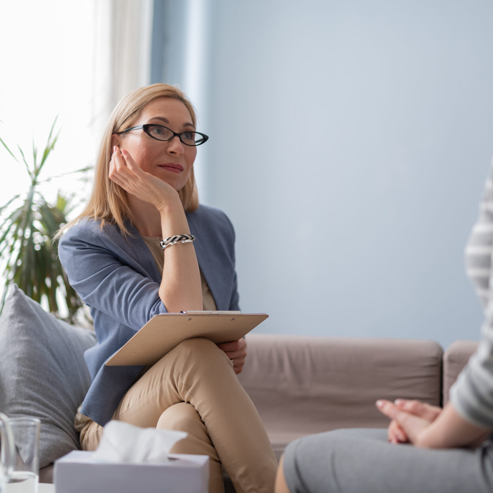 Psychotherapist in eyeglasses leaning on her elbow while looking at client and sitting on sofa in private consulting room. Brunette lady telling about traumatic events to a doctor indoors. Focus on coach